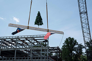 A steel construction beam suspended from a crane, covered in signatures, with a UVA flag and US flag hanging from it, topped with an artificial Christmas tree