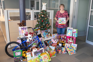 Brenda Buttner standing next to a Christmas tree surrounded by all the gifts donated by FM staff