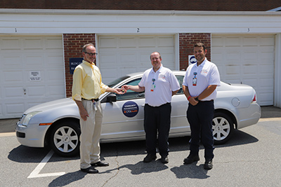 An FM employee hands a set of car keys to one of two paramedics standing in front of a Fleet vehicle branded with a Community Paramedicine - UVA Health door sticker