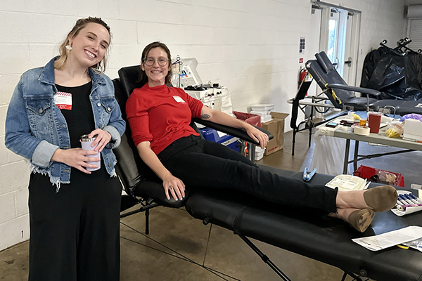 Katie Steele and Fiona Hogan smile at the camera, while Fiona is donating blood