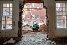 A mini-excavator in a courtyard, shown through a demolished brick and plaster wall