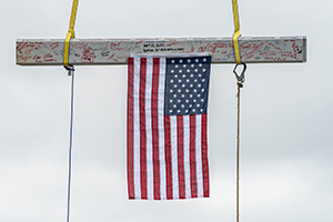 A steel beam suspended by a crane, covered in signatures and hanging an American flag