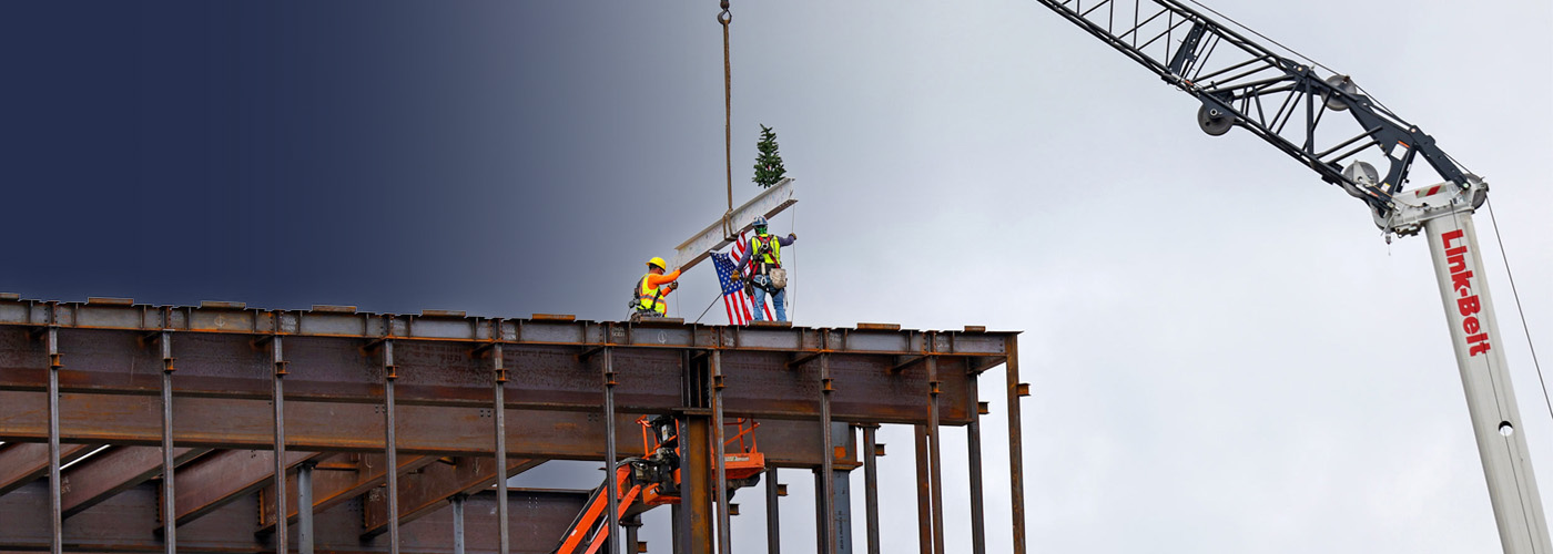 A construction girder being set in place by crane, with an American flag hanging from it and small pine tree perched atop it