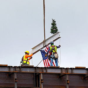 A construction girder being set in place by crane, with an American flag hanging from it and small pine tree perched atop it