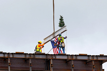 A construction girder being set in place by crane, with an American flag hanging from it and small pine tree perched atop it