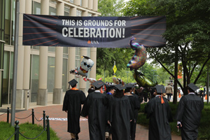 Students in caps and gowns walk beneath a banner reading 'This is Grounds for celebration!'