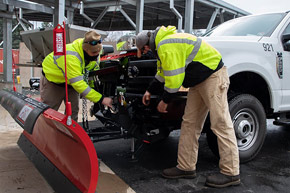 Two Facilities Management employees securing a snow plow to the front bumper of a pickup truck